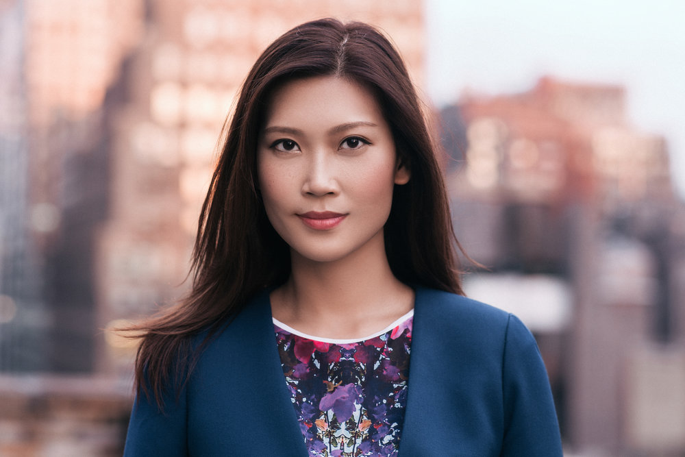 Headshot with a blurred city background of a stoic asian woman, wearing a blue business jacket, purple blouse with long straight hair.